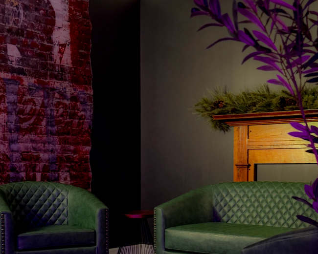 The image features two green velvet armchairs with a textured pattern, positioned against a brick wall and near a wooden mantel.