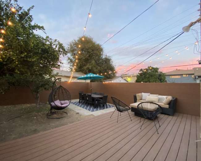 The image shows a backyard patio with a wooden deck, outdoor seating, a table under an umbrella, and string lights overhead.