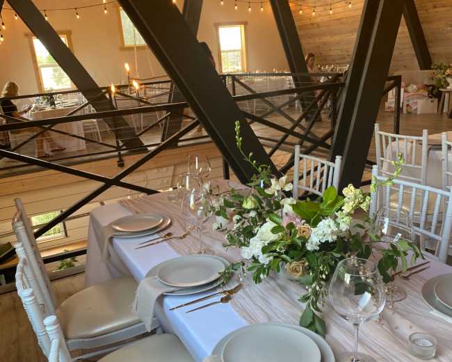 A dining table is elegantly set with plates, glasses, and floral arrangements in a rustic venue adorned with string lights.