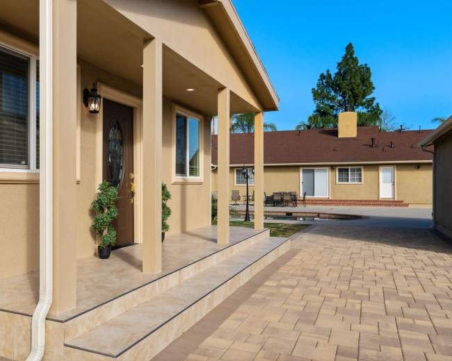 The image shows a house entrance with a covered porch, featuring a front door and potted plants, alongside a paved walkway leading to other buildings.