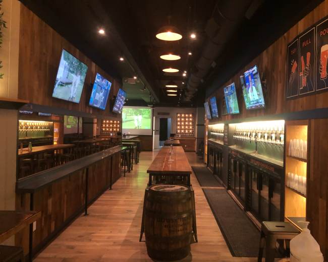 The image shows a modern bar interior with wooden walls, rows of taps, several flat-screen TVs displaying sports, and a long communal table.