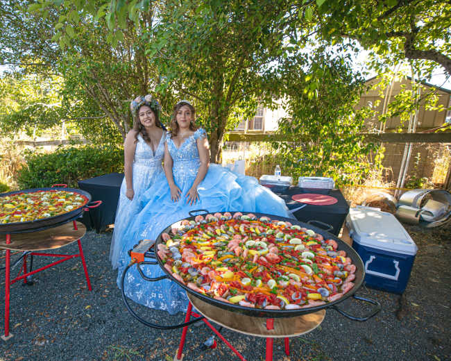 Two women in blue dresses stand beside large pans of colorful paella under a tree at an outdoor event.