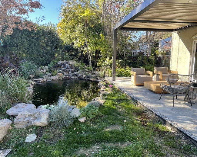 A patio area with wooden seating overlooks a pond surrounded by rocks and plants in a landscaped garden.
