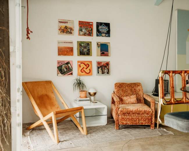 The image shows a cozy living space with two chairs, a small white table, and a wall adorned with colorful vinyl record covers.