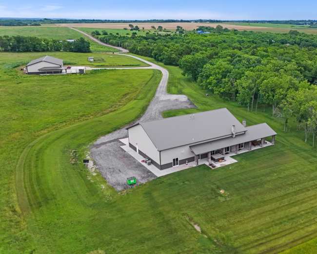The image shows a large, single-story building surrounded by grassy fields and trees, with a winding road leading to it from the background.