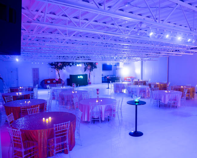 A spacious event venue with round tables set for dining, illuminated by purple and blue lighting.