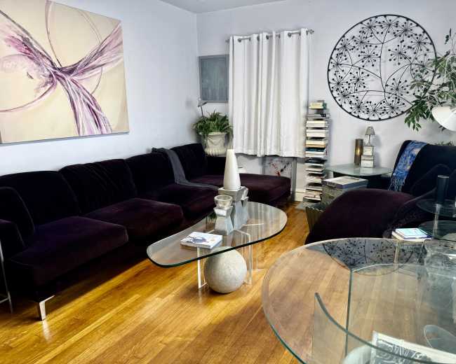 The image shows a cozy living room with a dark velvet sectional sofa, a round wall art piece, and glass tables atop wooden flooring.
