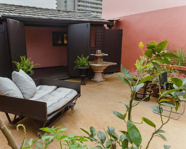A small outdoor area features a black wooden cart with cushions, surrounded by greenery and a stone pedestal.