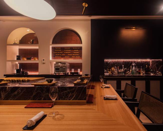 The image depicts a modern bar or dining area featuring a wooden countertop, elegant lighting, and shelves displaying glassware and various spirits.