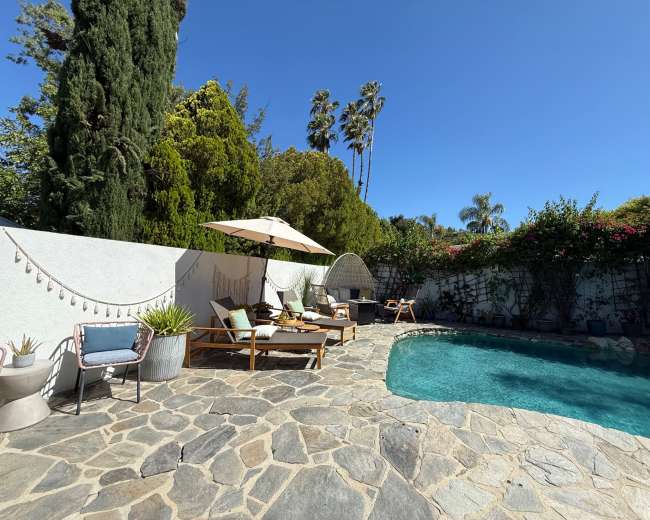The image shows a landscaped backyard with a swimming pool, stone patio, lounge chairs, and an umbrella surrounded by greenery and palm trees.