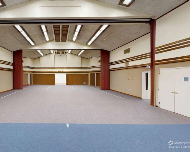 The image shows a large, empty interior space with a high ceiling, carpeted flooring, and several doors along the walls.