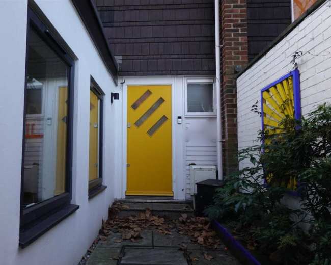 A narrow pathway leads to a bright yellow door flanked by white walls and surrounded by scattered leaves.