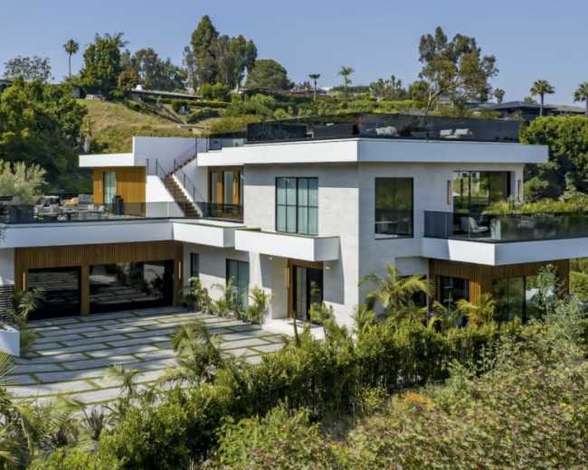 The image shows a modern two-story house surrounded by lush greenery and palm trees, with a stone pathway leading up to it.