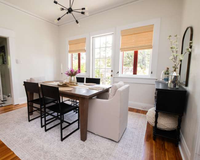 A dining room features a wooden table surrounded by black chairs, with large windows showcasing natural light and a rug on the wooden floor.