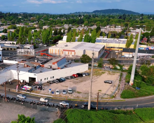 The image shows an aerial view of a mixed-use industrial area with various buildings, parked cars, and greenery in the background.