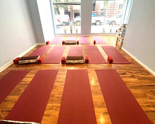 The image shows a yoga studio with red mats arranged neatly on a wooden floor, framed by large windows.