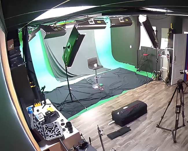 A video production studio is set up with a green screen, lighting equipment, and a camera on a tripod, while two individuals prepare for a shoot.