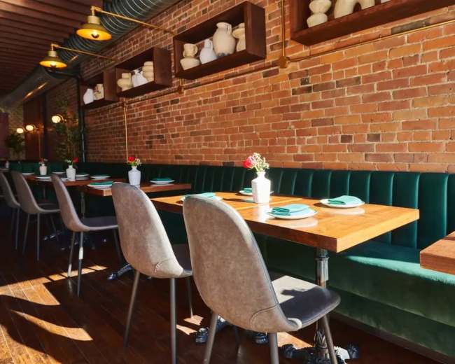 The image shows a restaurant interior featuring wooden tables, green upholstered seating, and brick walls adorned with decorative white ceramic vases.