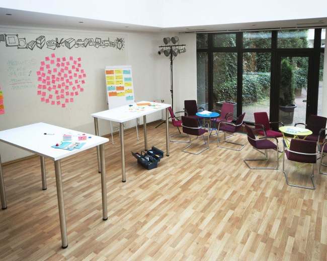 A spacious meeting room features several white tables, colorful sticky notes on the walls, and a cluster of chairs arranged around small tables.