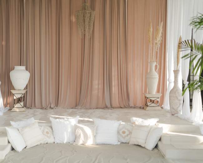 A stage is set with draped fabric in beige tones, flanked by large vases and cushions arranged on a sandy floor.
