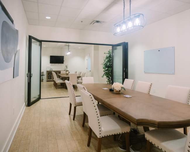 The image shows a formal conference room with a long wooden table surrounded by upholstered chairs, flanked by large glass doors leading to another meeting area.