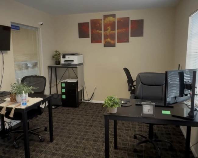 The image shows a small office space with two desks, computer monitors, a printer, and decorative wall art.