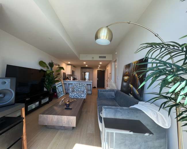 The image shows a modern living room featuring a large television, a sofa, a wooden coffee table, and a kitchen area in the background.
