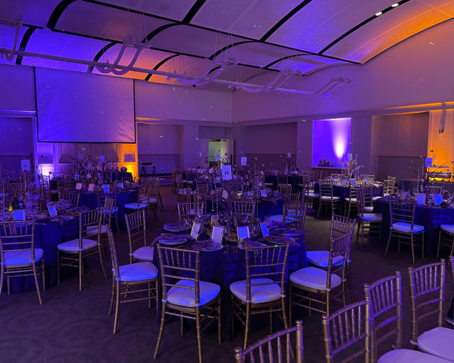 The image shows a banquet hall set up for an event, featuring round tables with blue tablecloths and gold chiavari chairs, illuminated by purple and orange lighting.