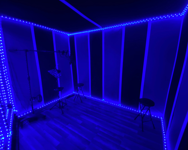 A sound recording studio with blue LED lights outlining the walls and two black stools positioned on a wooden floor.