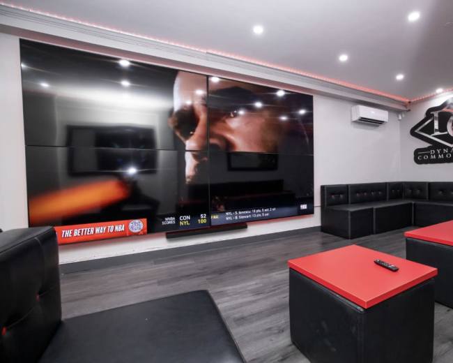 The image shows a modern entertainment room with large screens on the wall, black and red seating, and a low table in the center.