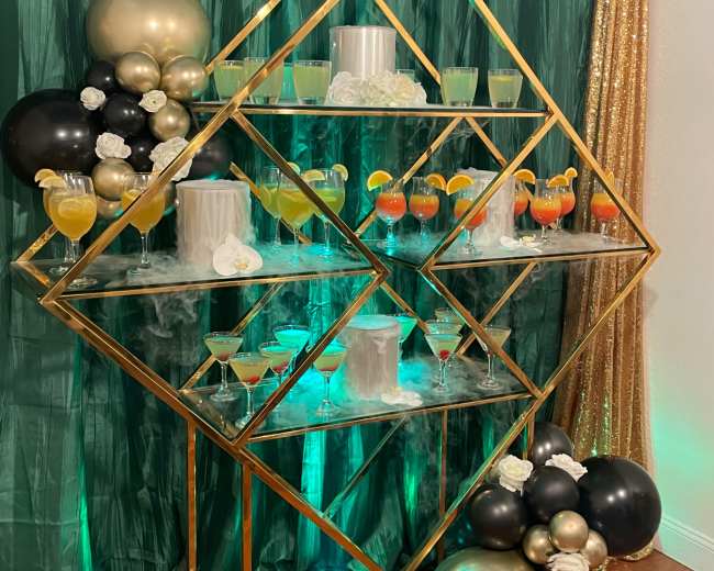 A decorative display features a geometric gold shelving unit adorned with various colorful drinks and balloons against a green and gold backdrop.