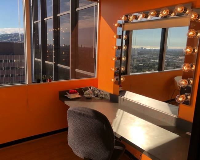 A brightly lit vanity area with a large mirror surrounded by bulbs, positioned near a window with a view of the city.