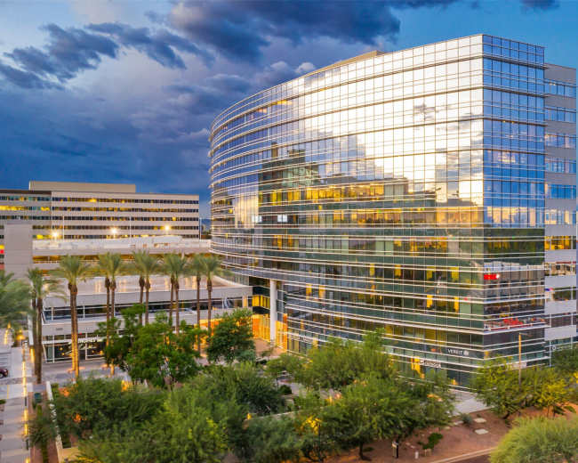 The image shows a modern glass office building surrounded by palm trees and additional structures under a cloudy sky.