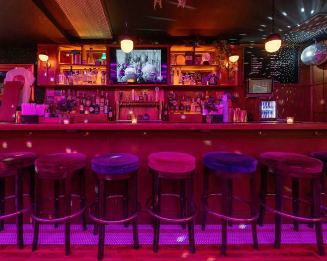 The image shows a dimly lit bar with a long counter, high stools, and shelves filled with various bottles, illuminated by colorful lights.
