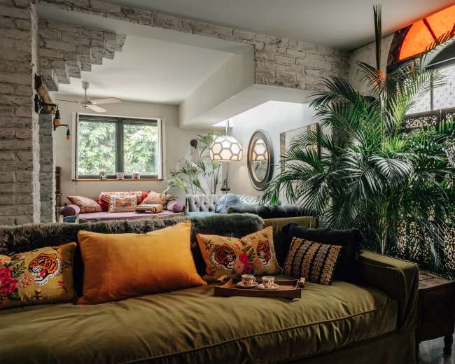 A spacious living room features a green couch adorned with colorful cushions and a wooden tray, surrounded by large plants and bathed in natural light.