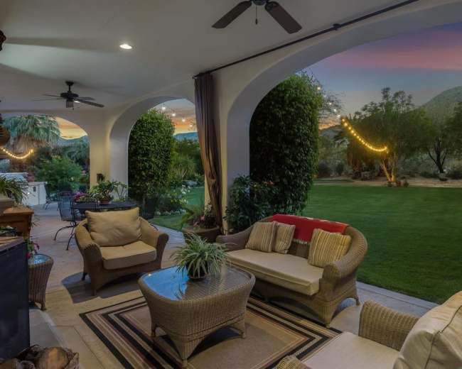 A spacious patio features comfortable seating and a view of a well-maintained lawn framed by trees, with hanging lights illuminating the scene at dusk.