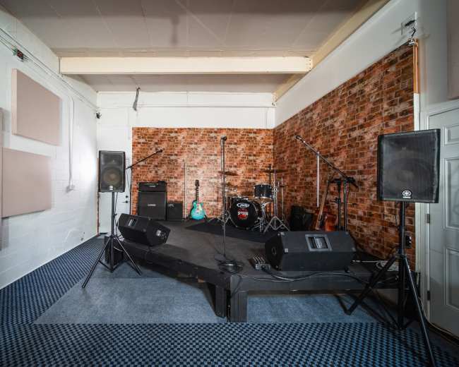 The image shows a small stage setup in a room with a brick wall, equipped with speakers, microphones, and musical instruments.
