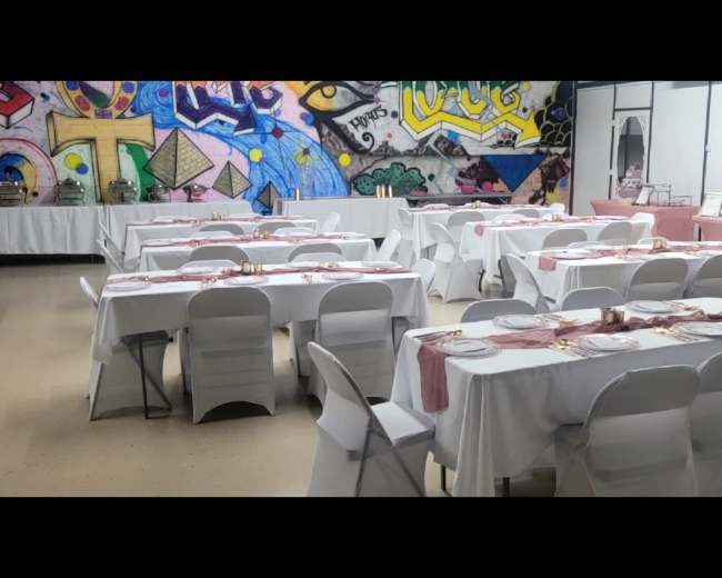The image shows a banquet hall with several white tables covered in tablecloths and set with dinnerware, against a backdrop of colorful graffiti art on the wall.