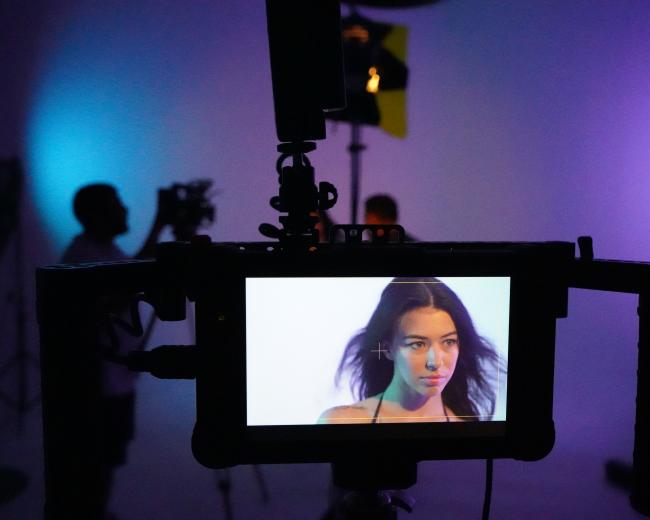 A camera monitor displays a close-up of a woman with flowing hair against a softly lit background, while crew members work behind her.