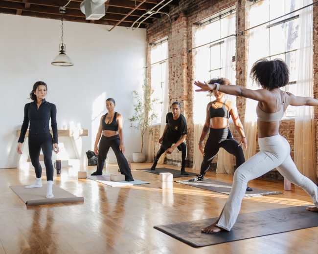 A group of five people practice yoga in a spacious, sunlit studio with brick walls and wooden floors.