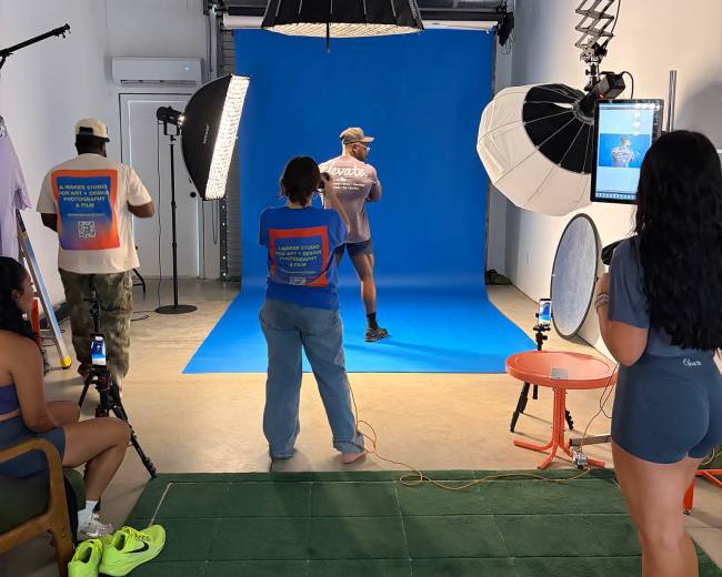 A photography studio features a model posing against a bright blue backdrop, surrounded by photographers and lighting equipment.