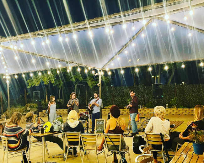 A small crowd watches a live music performance under a lit tent with string lights.