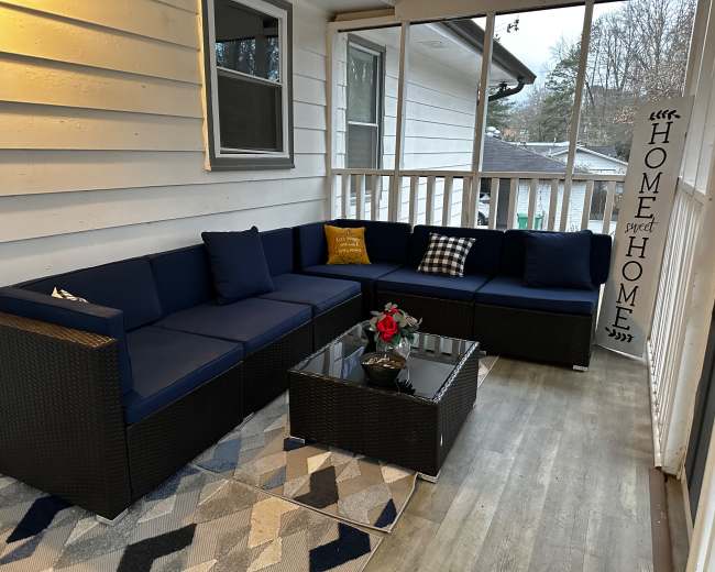 The image shows a porch area furnished with a sectional couch, a glass coffee table, and decorative items, all set against a backdrop of a white wall.