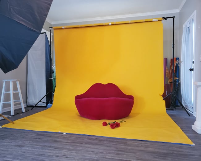 A red lip-shaped sofa sits in front of a bright yellow backdrop, accompanied by three red roses, in a studio setting.