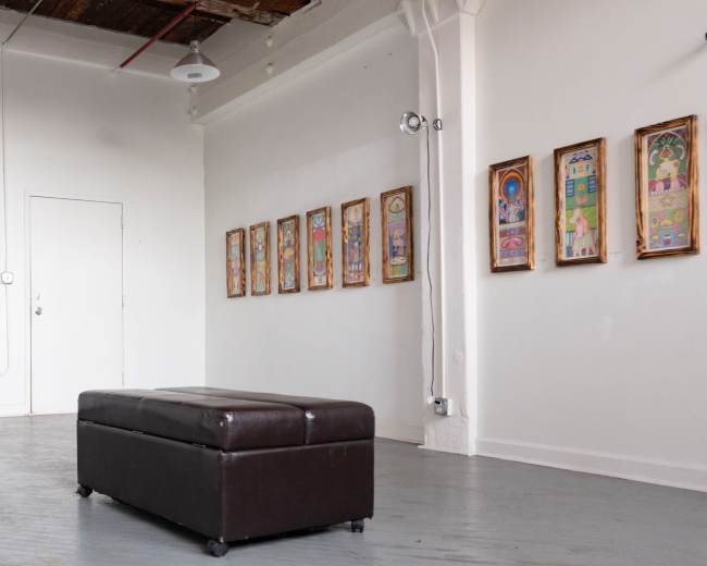 The image shows an art gallery with framed artworks hanging on the wall, a brown ottoman in the center, and a white door in the background.