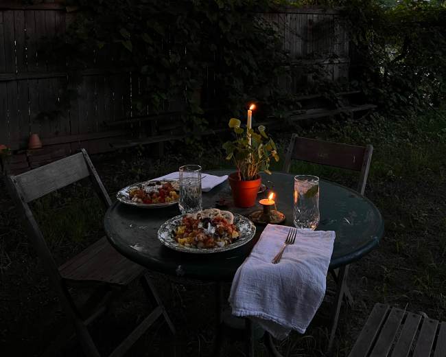 A small round table is set for two with plates of food, glasses of water, and a candle in a potted plant, beneath string lights illuminating a backyard at dusk.