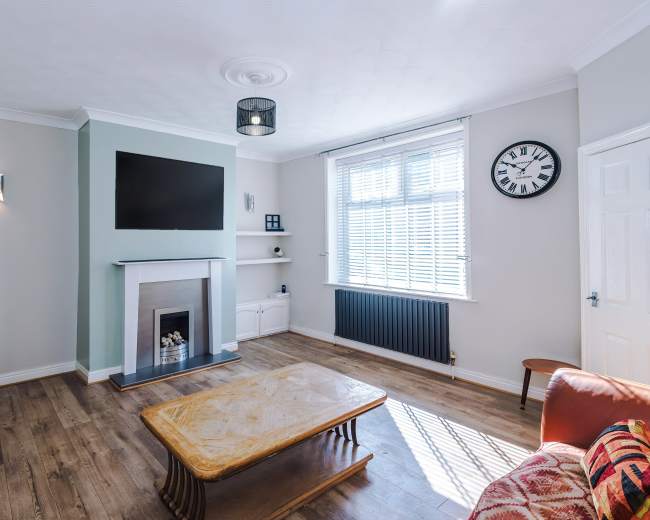 The room features a modern living area with a wooden coffee table, a flat-screen TV mounted above a fireplace, and large windows allowing natural light.