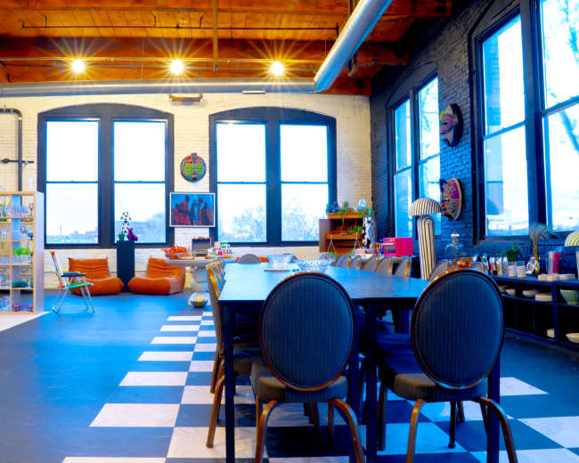 The image depicts a spacious room with large windows, wooden beams, a long dining table, colorful seating, and various decorative elements.