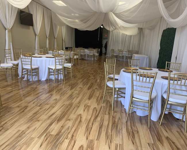 The image shows a banquet hall with several tables dressed in white tablecloths and gold chairs, featuring a wooden floor and draped fabric overhead.