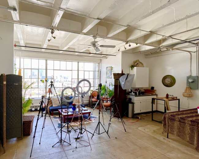 A spacious room with large windows features various photography equipment, green plants, and furniture arranged for a creative setup.